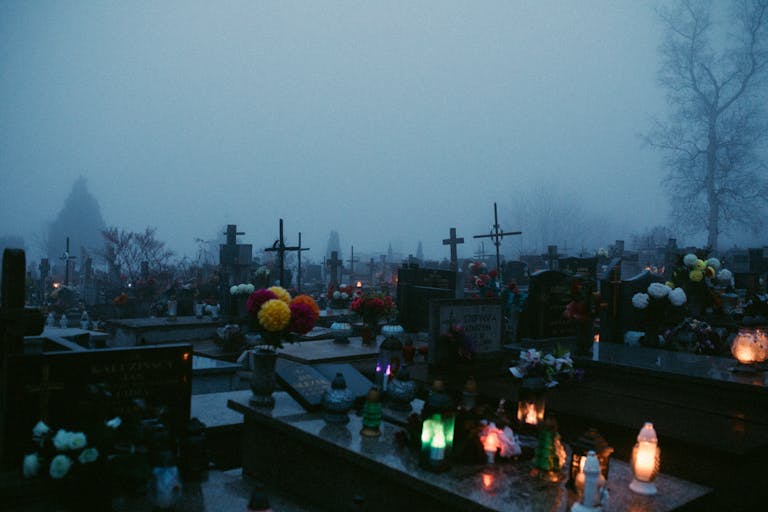 A foggy cemetery at night illuminated by candlelights on the graves, evoking a somber ambiance.