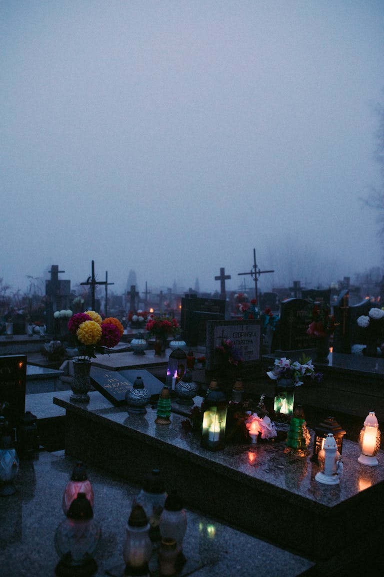 A misty and solemn cemetery scene with illuminated gravestones and flowers, evoking a reflective mood.