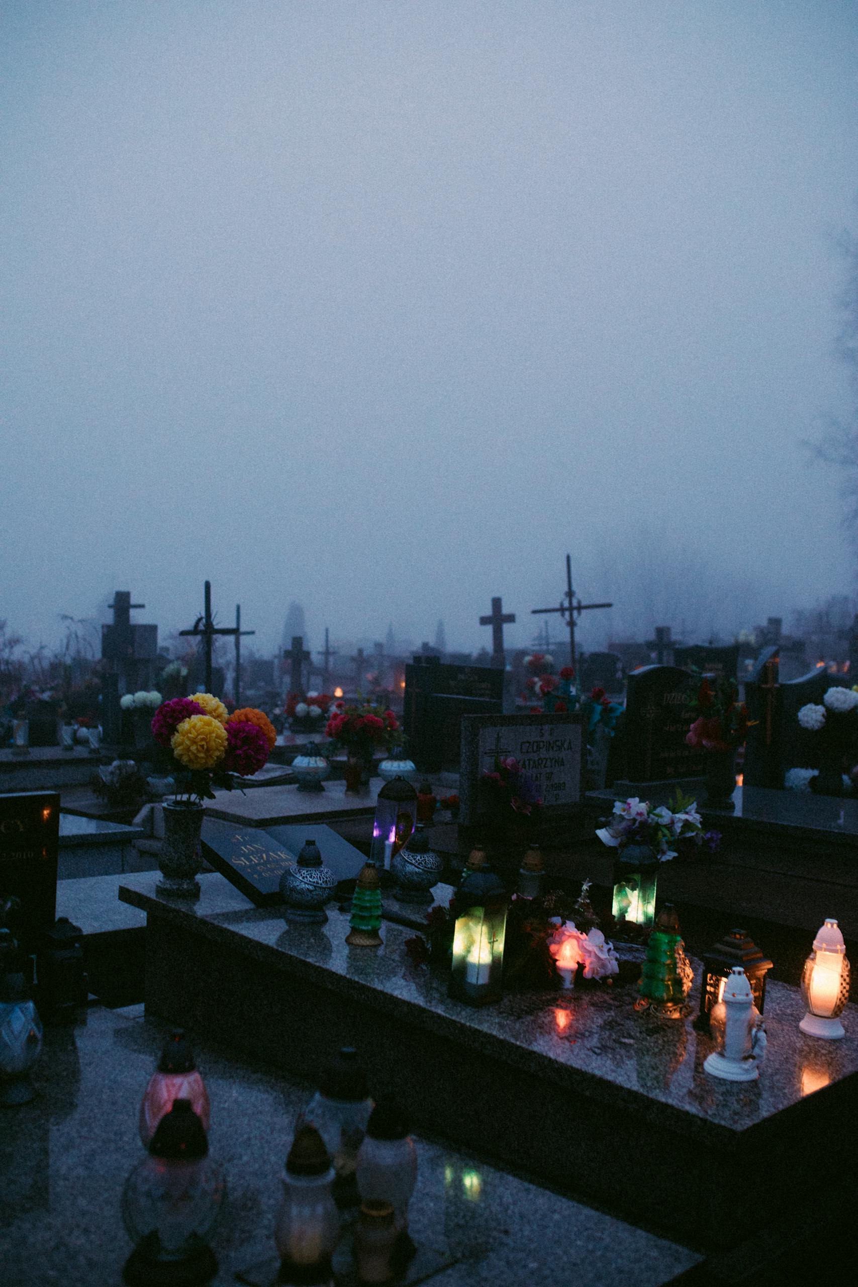 A misty and solemn cemetery scene with illuminated gravestones and flowers, evoking a reflective mood.