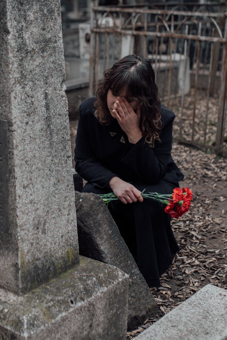 A sad woman kneels by a tombstone in a cemetery, holding red flowers in mourning.